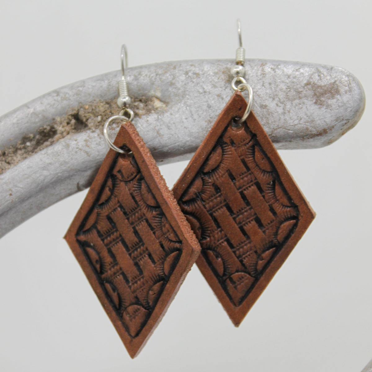 A pair of handmade wooden diamond-shaped dangle earrings with carved basketweave patterns, hanging from silver fishhook wires on a rustic curved branch prop against a light gray background.